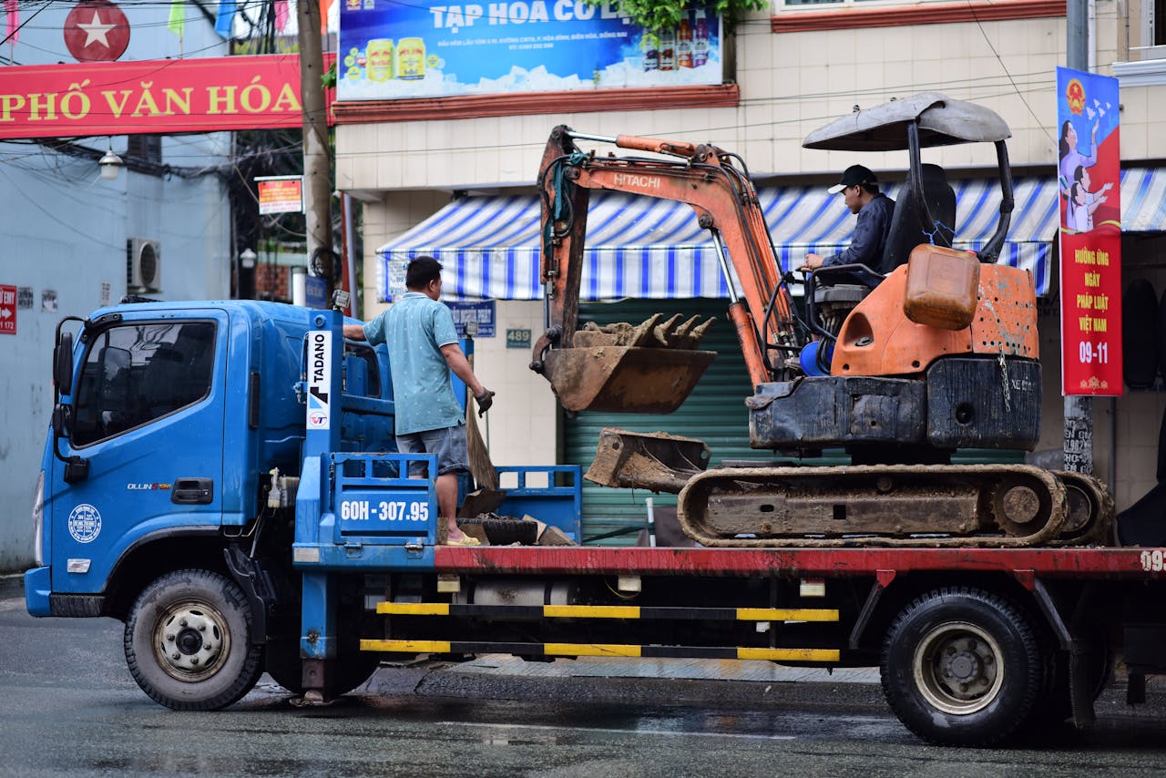 الرئيسية Two men and excavator being transported by truck on rainy urban street.